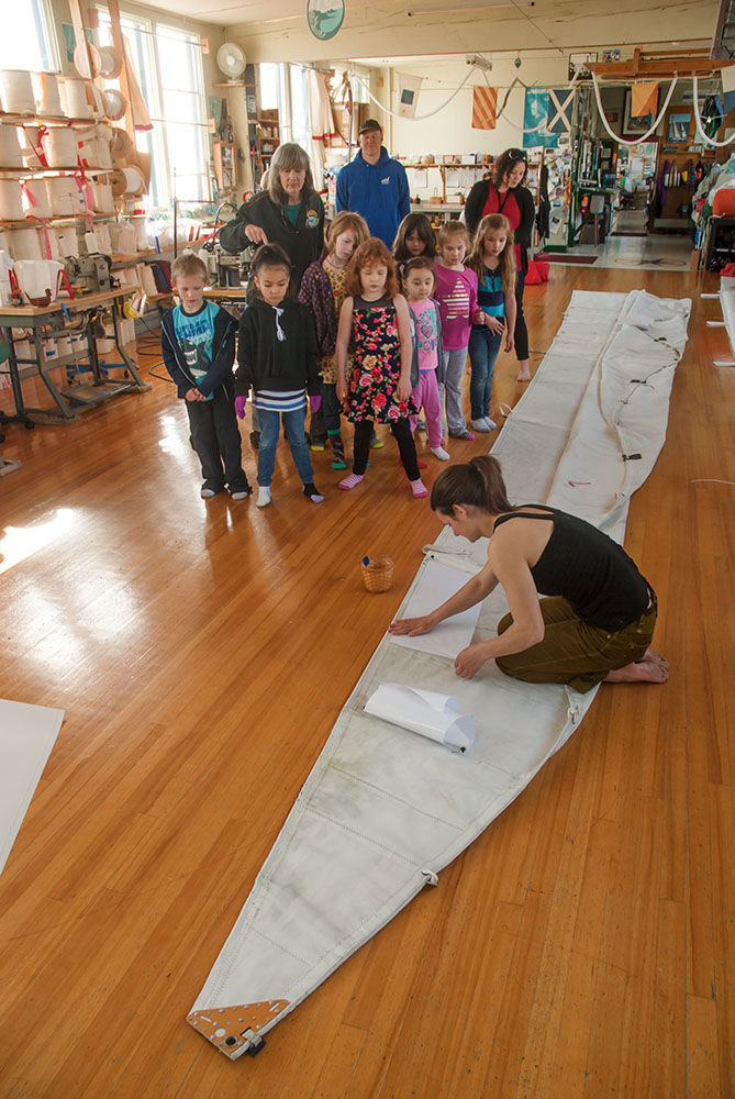 “Sails are wings,” says Carol Hasse, owner of Port Townsend Sails (standing behind children, at left, in black jacket) as she shows a group of Grant Street Elementary first-graders a sail being repaired on April 11. Pictured (from left) are Tarrence W., Nyla R., Carol Hasse, Malia W., Hope Q., Naveah G., Sophia B., Lily R., Idalya S. and Ashley Quinn. Working on the sail is employee Erica Georgaklis. Photo by Chris Tucker