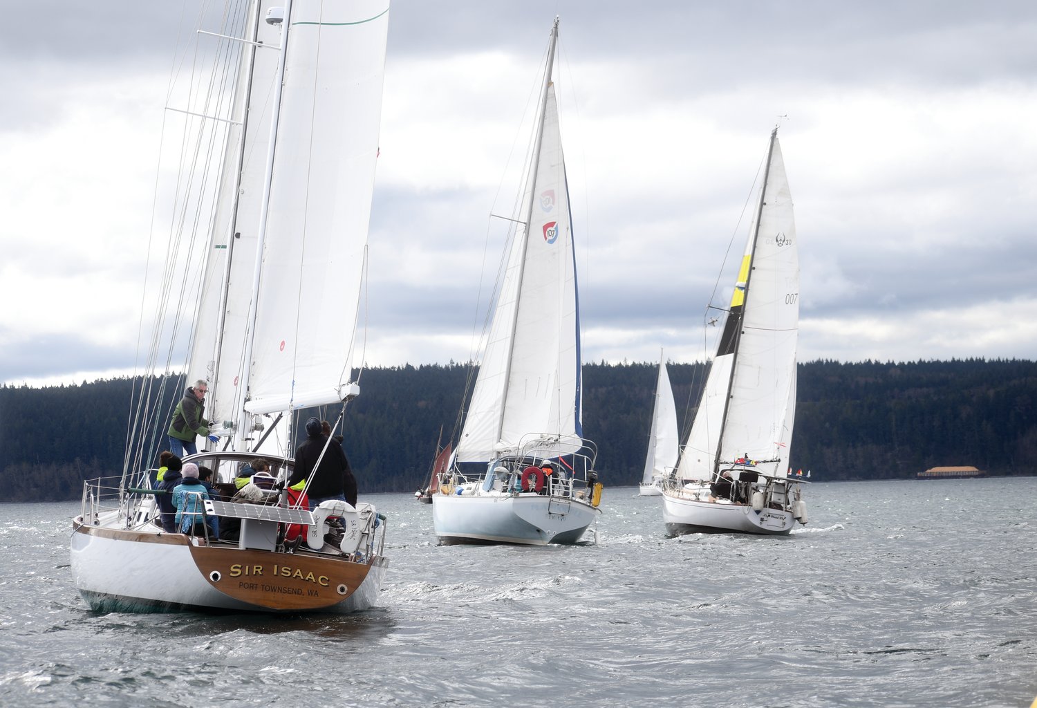 Sailors from last year’s Shipwrights’ Regatta glide toward Boat Haven Marina through Port Townsend Bay, with the local landscape behind them.